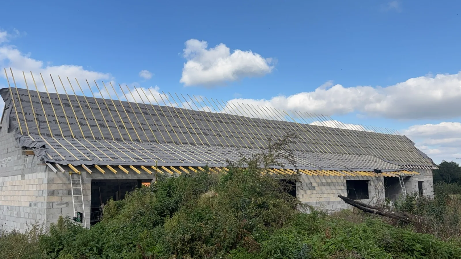 Grand bâtiment agricole en parpaings avec couverture neuve en plaques grises, pannes visibles, ciel nuageux